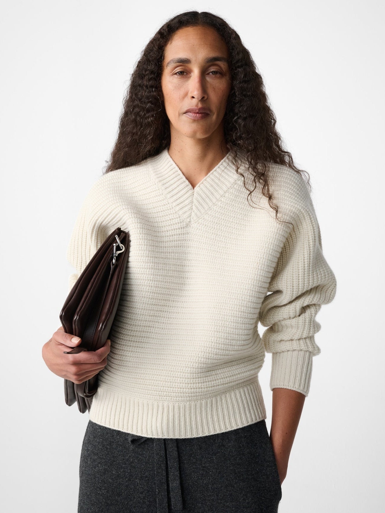 A model with long curly dark hair wearing a cream ribbed knit V-neck sweater, dark gray drawstring trousers, and holding a dark brown leather clutch against a light gray studio background.