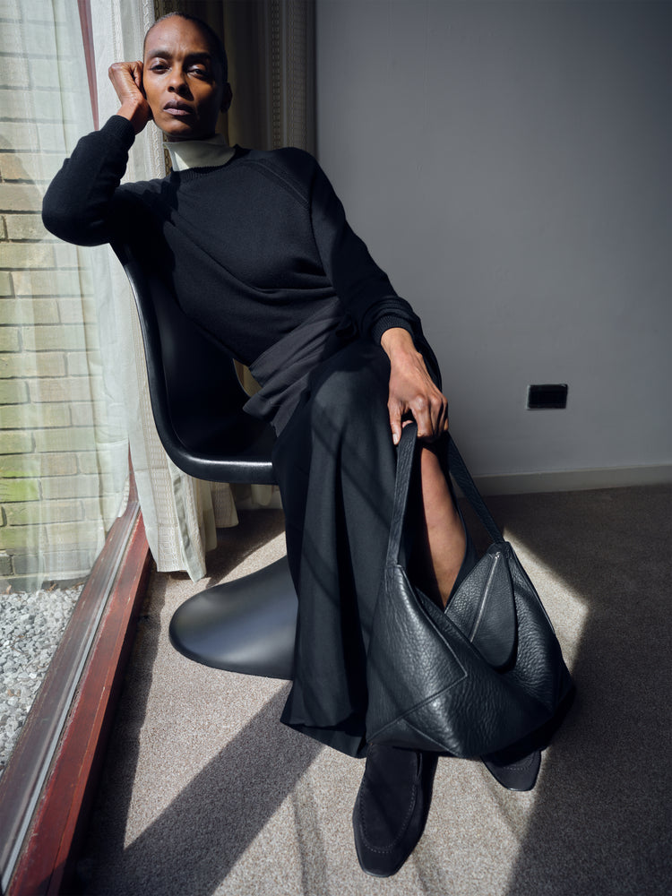 Woman holding a black leather bag in a room with a window and gray wall wearing a black skirt.